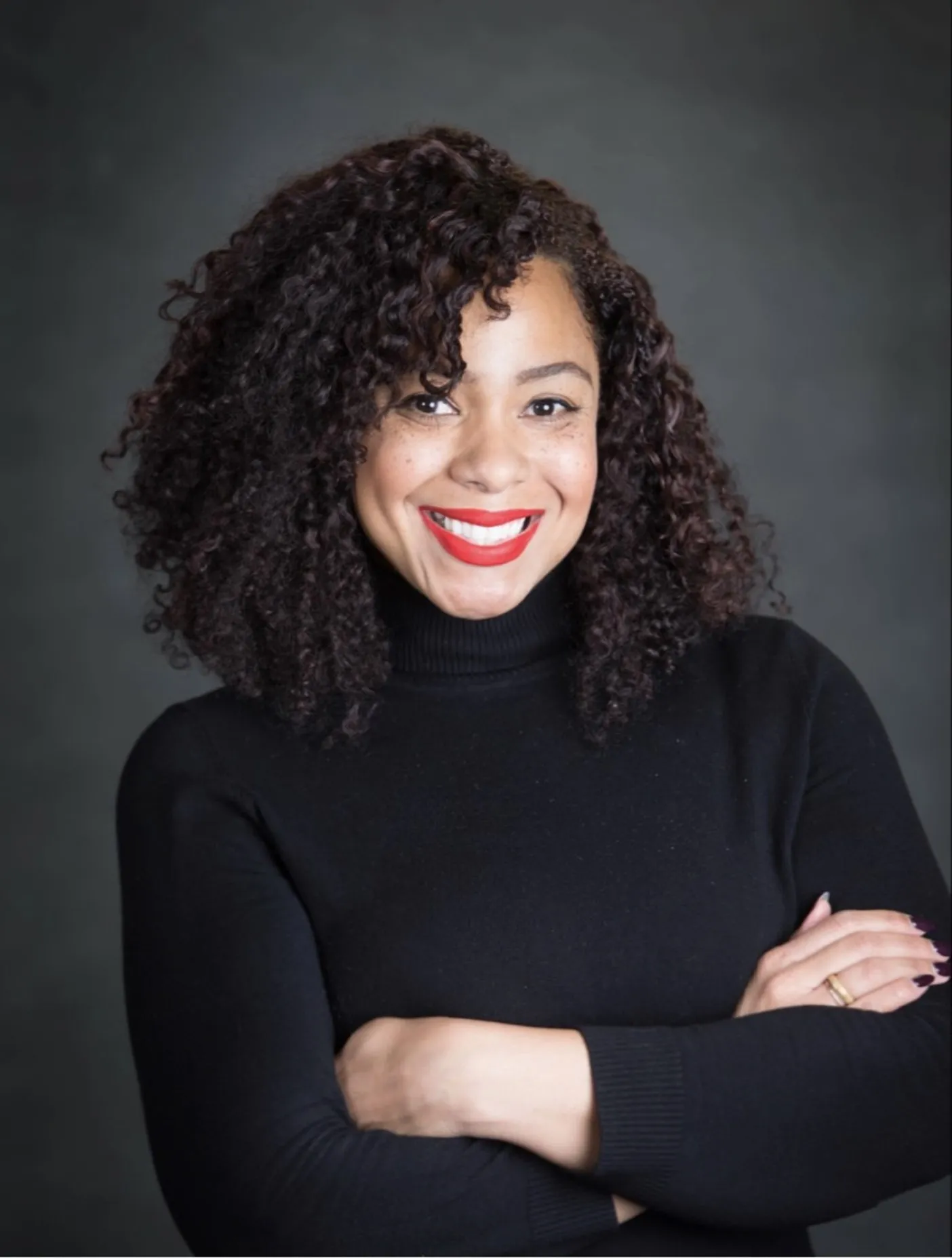 Smiling woman with curly hair wearing a black turtleneck sweater and red lipstick, arms crossed.